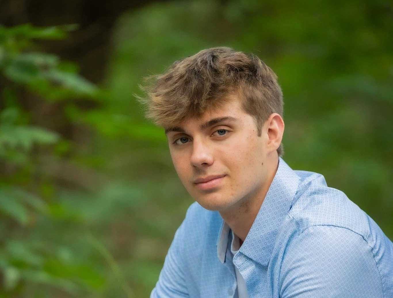 Young man in a blue shirt posing outdoors with a backdrop of lush green foliage.