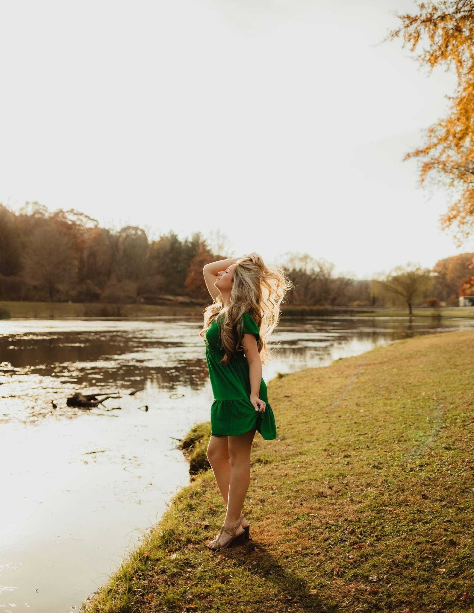 Woman in a green dress standing by a lake in a park, with autumn trees in the background.